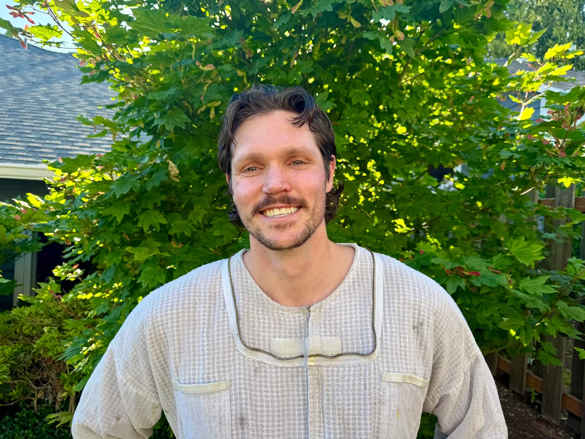 Sam, who runs Sam The Bee Man on his own, smiling in a cream bee jacket in a sunny backyard